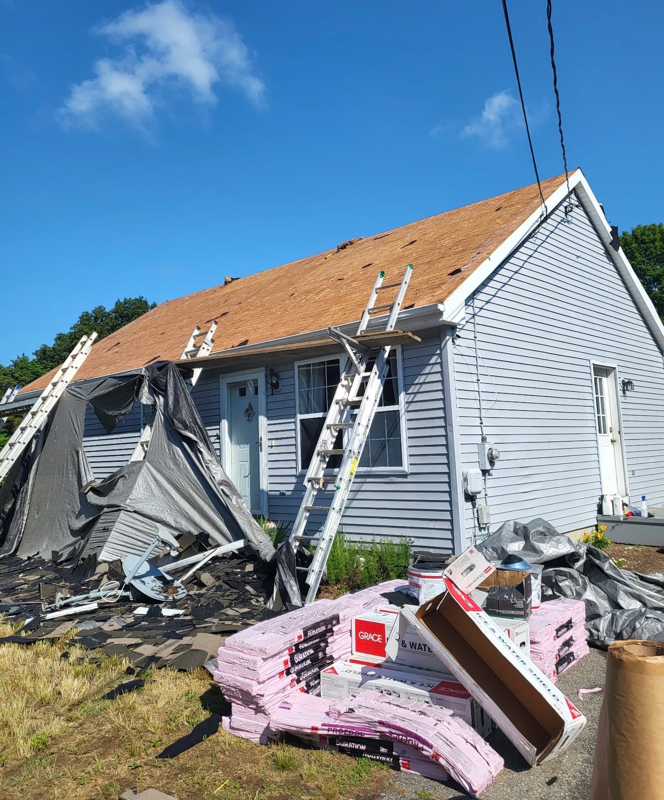 Blue house undergoing roof replacement with ladders, exposed plywood, and piles of roofing materials.